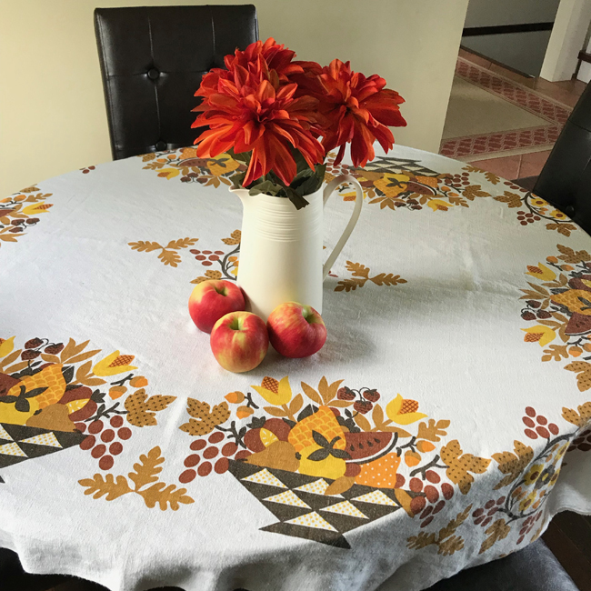 Printed tablecloth with orange and brown leaf es, and a white pitcher filled with orange flowers