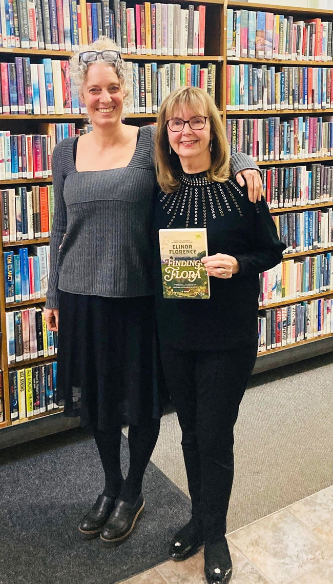 Two women, one blonde and one brunette, stand together in front of wall of bookshelves.