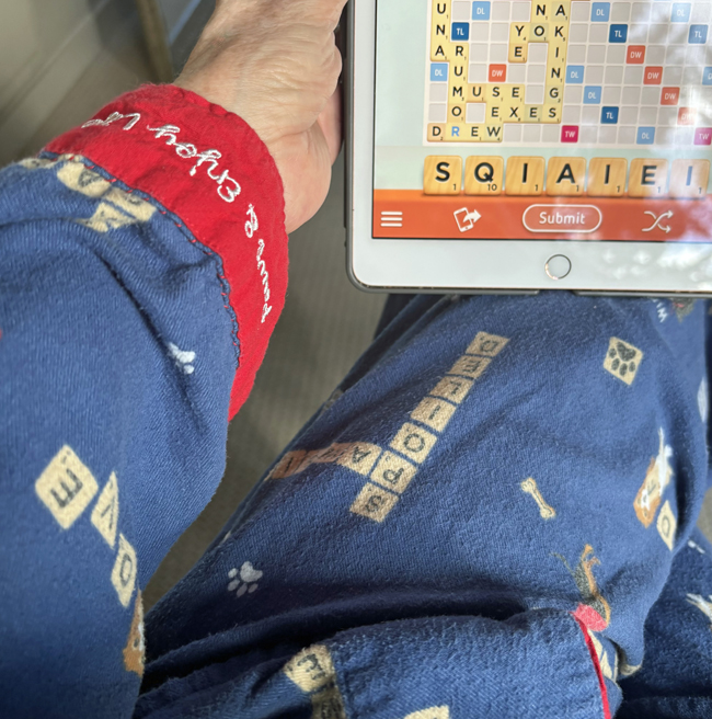 Woman wearing red and blue pyjamas printed with scrabble words holds iPad in one hand showing scrabble game