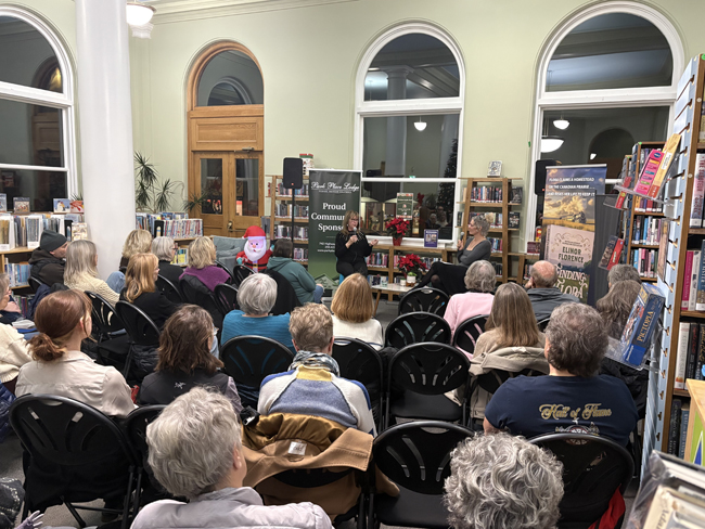 Audience seated in front of two women at the front, sitting under three magnificent arched windows.