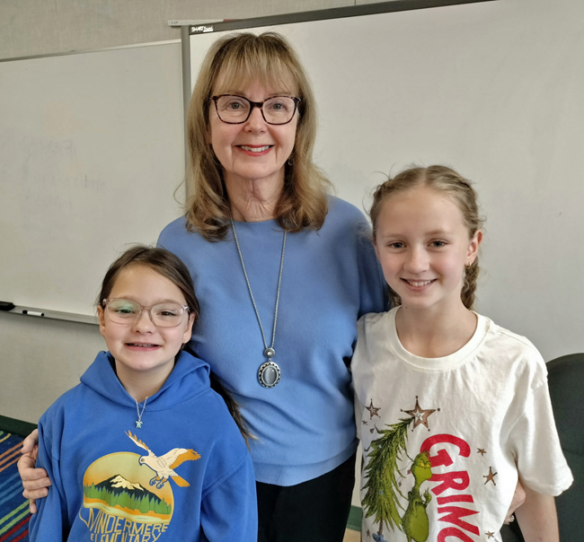 Smiling woman in blue sweater stands between two little girls, one blonde and one brunette.