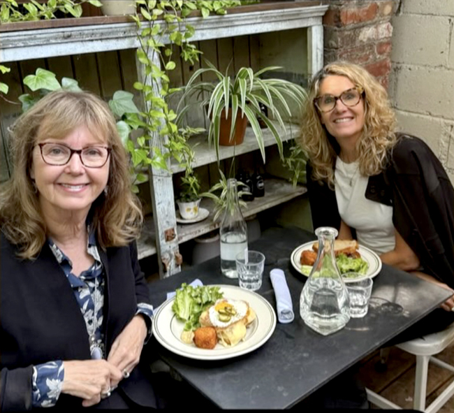Two smiling women, one blonde and one brunette, both wearing glasses, seated at a lunch table