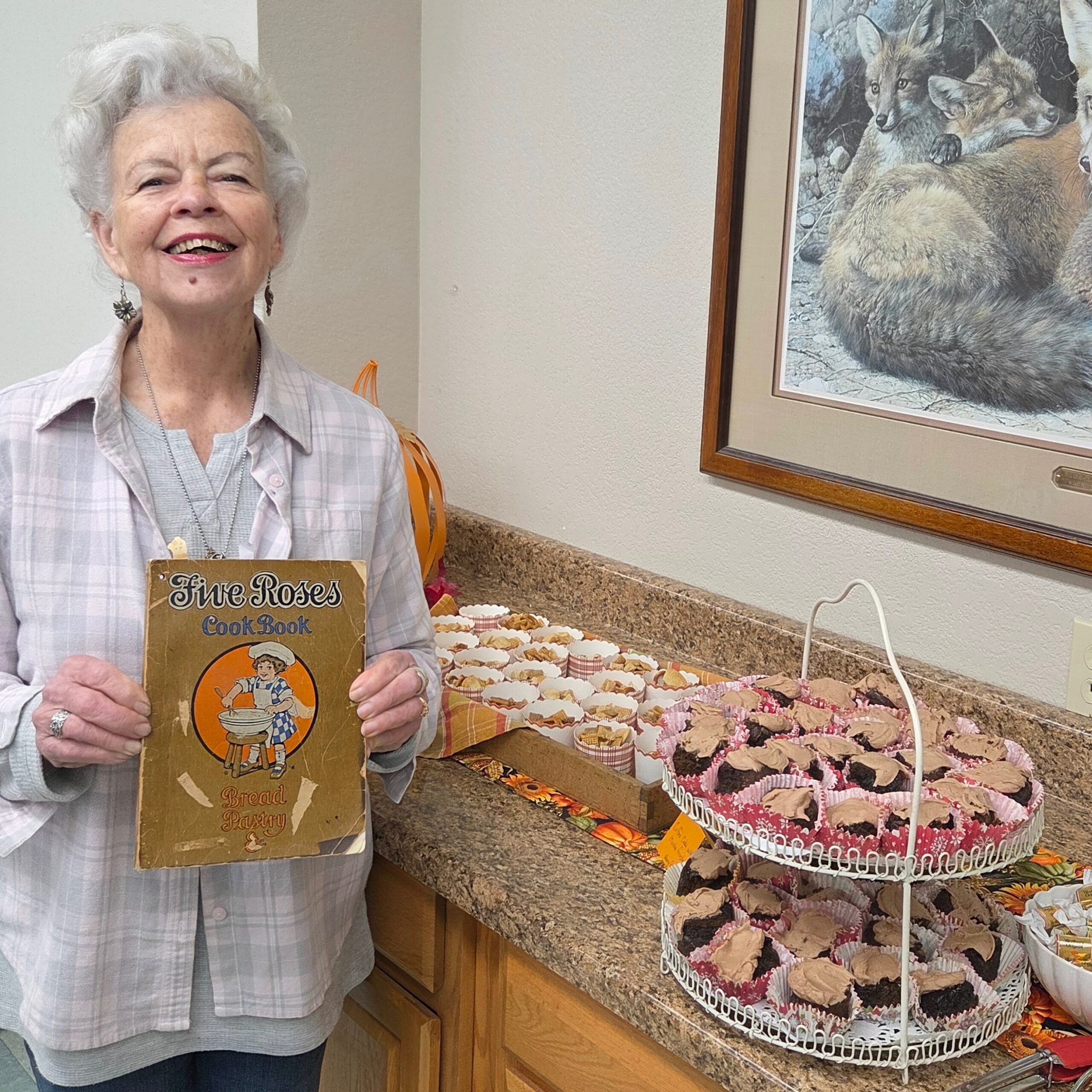 Smiling white-haired woman holding cookbook stands beside cake stand filled with muffins